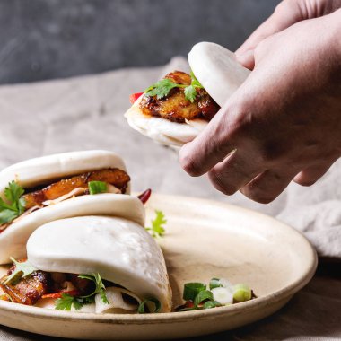 Adamın elleri Asya sandviç buğulama gua bao buns domuz eti, yeşil ve sebzeler ile keten masa örtüsü seramik tabak masaya servis ile tutun. Asya tarzı hızlı yiyecek yemek. Kare görüntü
