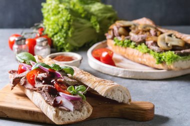 Gri mavi masa üzerinde malzemelerle seramik tabak etli baget sandviç domates, fesleğen, kırmızı soğan ile servis.