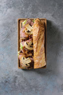 Sığır eti baget sandviç champignon mantar, yeşil salata, kızarmış soğan ile gri doku arka plan üzerinde malzemelerle ahşap hizmet gemide görev yaptı. Düz lay, uzay