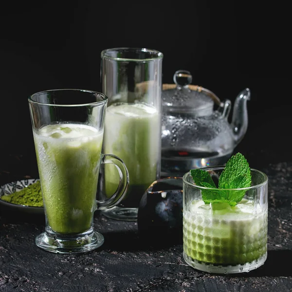 Matcha green tea iced latte or cocktail in three different glasses with ice cubes, matcha powder and transparent teapot over dark texture background. Square image