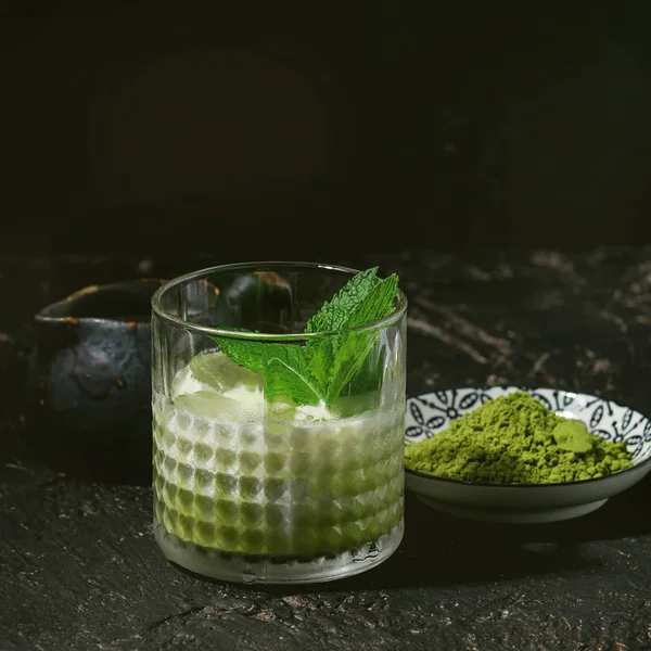 Matcha green tea iced latte or cocktail in glass with ice cubes, mint, matcha powder and jug of milk over dark texture background. Square image