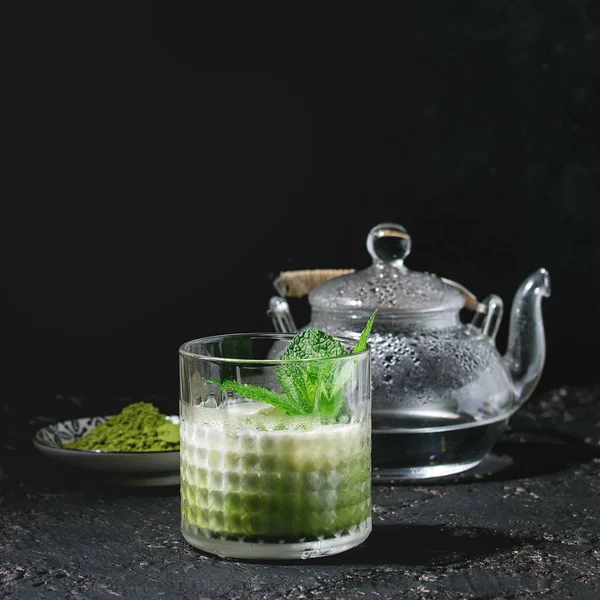 Matcha green tea iced latte or cocktail in glass with ice cubes, mint, matcha powder and transparent teapot with hot water over dark texture background. Square image