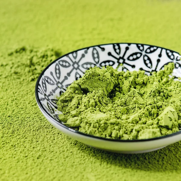 Green tea matcha powder in ceramic bowl over powdering matcha as background. Close up. Square image