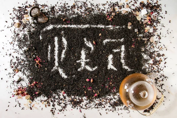 Calligraphic inscription gothic letters tea over dry black ceylon and green tea scattered on white marble with rose buds, stainer, glass teapot. Tea drinking concept background. Top view