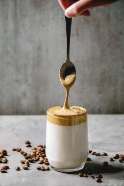 Dalgona frothy coffee trend korean drink milk latte with coffee foam in glass mug, foam flowing from spoon. Gray texture table. Copy space clipart