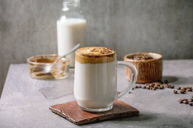 Dalgona köpüklü kahve trend Kore sütlü latte, cam bardakta kahve köpüğü ve gri dokuma masasında öğütülmüş kahve ile süslenir. Yukarıdaki malzemeler. Boşluğu kopyala
