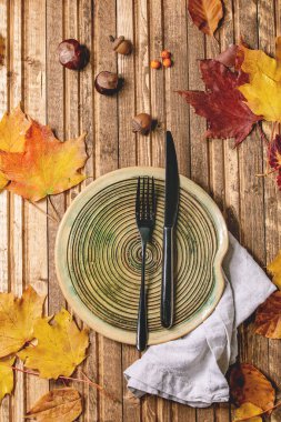 Ahşap zemin üzerinde kırmızı ve sarı sonbahar yaprakları üzerinde tekstil peçetesi ve çatal bıçak bulunan boş seramik tabaklar. Düz yatıyordu. Sonbahar yemekleri arka planı.