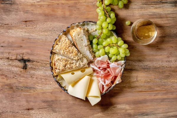 Appetizers antipasti with white sicilian focaccia. Traditional bread sliced cake with onion served with prosciutto ham, cheese, grapes and glass of white wine over wooden background. Flat lay, space