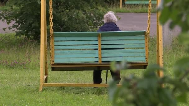 在秋阴天，一位老妇人坐在花园秋千上秋千. — 图库视频影像