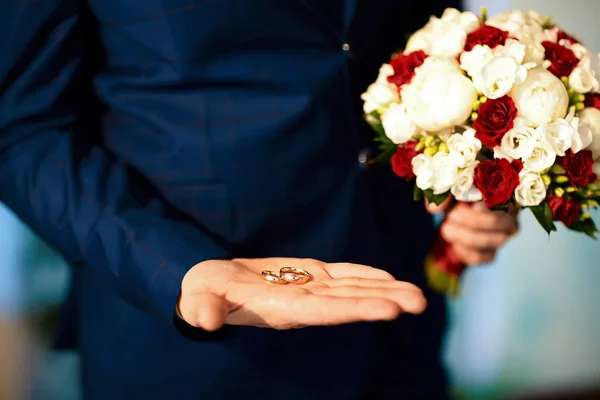 花嫁の手の中の金の結婚指輪白と赤の花の花束とスーツ — ストック写真