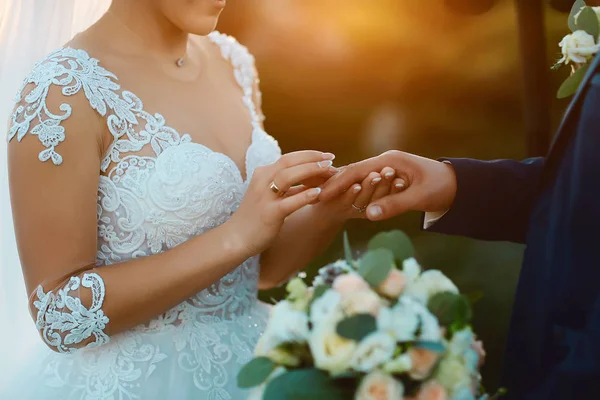 日没の美しい結婚式 白いエレガントなドレスの花嫁は手を保持し 暗いスーツで彼女のスタイリッシュな新郎にリングを屋外に置きます ウェディングウェア アクセサリー — ストック写真