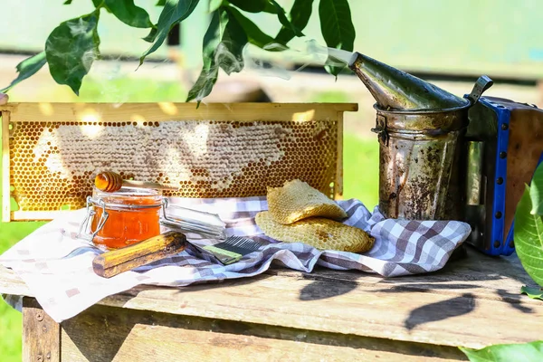 Jar of fresh honey with assorted tools for beekeeping, a wooden dispenser and tray of honeycomb from a bee hive in a still life on a wooden table outdoors.
