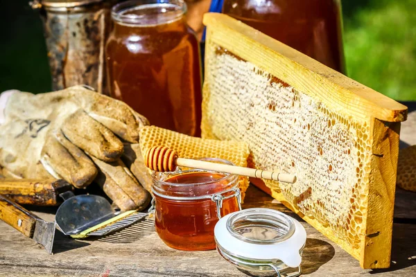 En burk färsk honung med olika verktyg för biodling, en trägolv och en bikupa i ett stilleben på ett träbord utomhus med kopieringsutrymme — Stockfoto