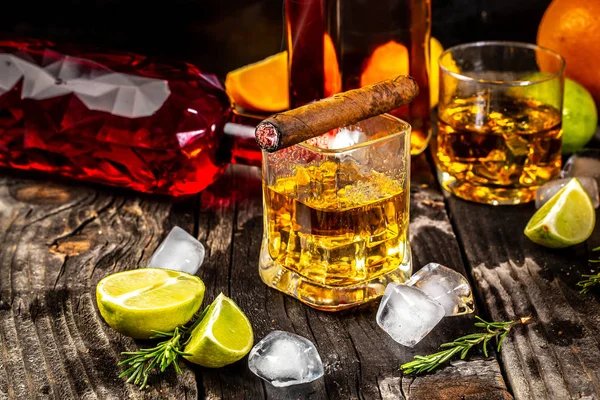 Vaso de whisky con cigarro ahumado y cubitos de hielo sobre mesa de madera. Bebida alcohólica diferente en vaso. concepto bebida de lujo — Foto de Stock