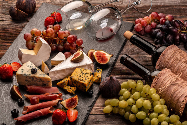 still life, food composition with assorted cheese, glass of wine and fruits on a wooden table.