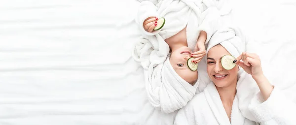 Positive mother and daughter doing face mask applying pieces of cucumber to their eyes, wrapped in a white bath towel, Facial skin care, cosmetology and spa, Long banner format. space for text.