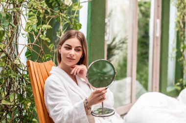 Genç ve güzel bir kadın yüzüne dokunuyor ve aynaya bakıyor. Güzellik, cilt bakımı ve insanlar. Sağlık ve güzellik, tatil beldesi ve rahatlama konsepti.