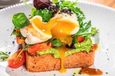 Avokado tostlu haşlanmış yumurta, vişneli domates, taze yeşiller ve beyaz tabakta pişmiş yumurta sarısı..