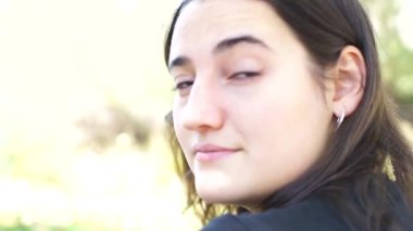 Uzun kahverengi saçlı genç beyaz kadın döner ve ön plandaki kameraya sakince bakar.