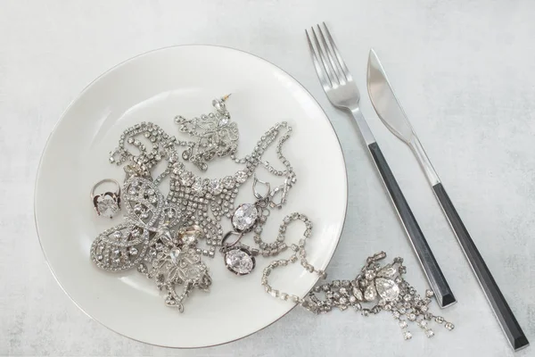 Many sparkling fake diamond jewelery on a plate with a knife and fork. The concept of luxury life, wealth, glamor, fashion and weddings