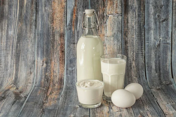 Latte in una bottiglia uova panna acida e un bicchiere di latte su uno sfondo di legno — Foto Stock