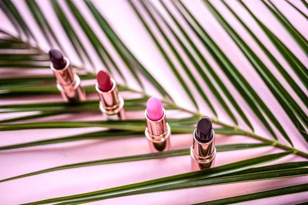 Multicolored lipsticks in metal tubes on a pink background on a sheet of palm trees with selective focus — Stock Photo, Image