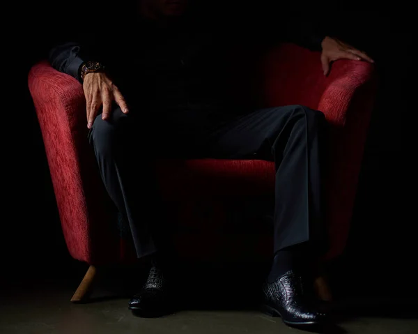 male businessman in a black suit sitting in red chair, black background, no faces visible, studio shooting