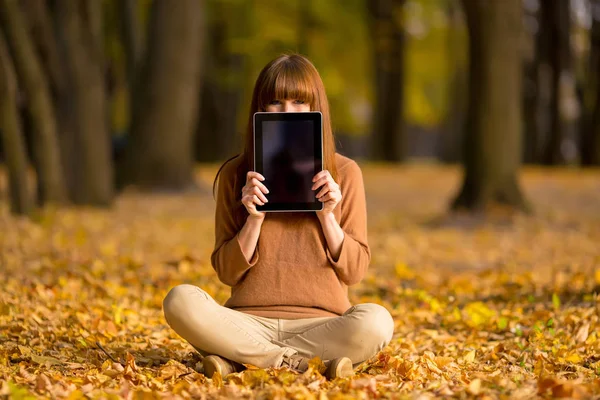 smiling young woman showing tablet pc sitting on fallen leaves in autumn park