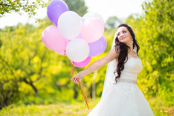 Noiva deslumbrante feliz no vestido com balões coloridos no parque verde — Fotografia de Stock