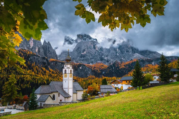 Amazing autumn scenery with colorful larch forest. Charming alpine village with beautiful church. Great hiking and touristic places, Colfosco, Dolomites, Italy, Europe