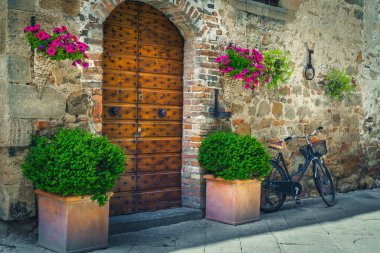 Tahta kapılı tuğla kemerli giriş. Çiçek saksısı ve renkli çiçeklerle süslenmiş giriş. Giriş ve park edilmiş bisikletli sokak manzarası, Pienza, Toskana, İtalya, Avrupa