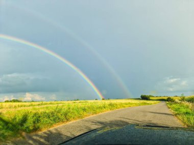 Yeşil bir çayırda çifte gökkuşağı manzarası ve yaz yağmurundan sonra bir arabadan görülen köy yolu.