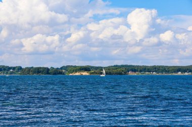 Beyaz yelkenli, kumlu uçurumlarla dolu ormanlık kıyı şeridi boyunca derin mavi sularda süzülüyor. Kabarık bulutlar, sakin deniz sahnesinde parlak yaz gökyüzünü noktalıyor.