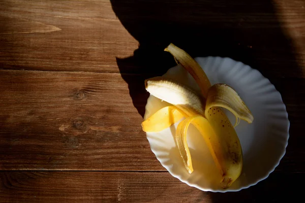 在被太阳照亮的白色盘子上去皮的香蕉 — 图库照片