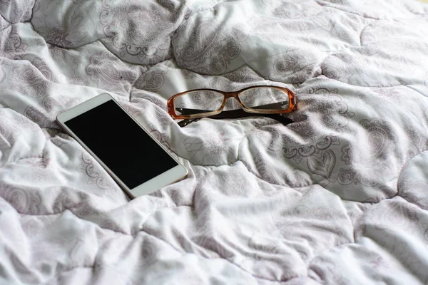 on a crumpled bed are glasses and a white smartphone. Lighting - light from the window. Concept - morning mess. The begining of the work day.