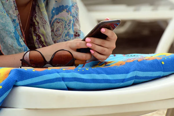 Plaj şezlongundaki bir kadın elinde bir akıllı telefon tutuyor. Yakındaki güneş gözlüğü. Konsept - plaj tatili