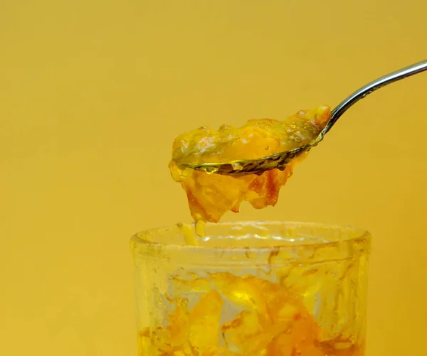 A teaspoon is filled with orange jam. A spoon is located above a glass of orange jam. Yellow background. The concept is sweets. Drink tea with jam