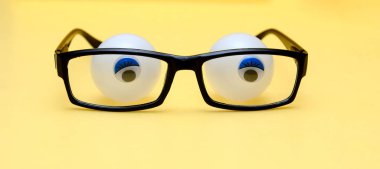  Sarı arka plandaki siyah gözlük çerçevesi. Gözlüklerin arkasında yapıştırılmış gözbebekleri olan tenis topları var. Kavram - gözlük gözlük arkasında gözler, korku, karışıklık, düşüncelilik ifade. 