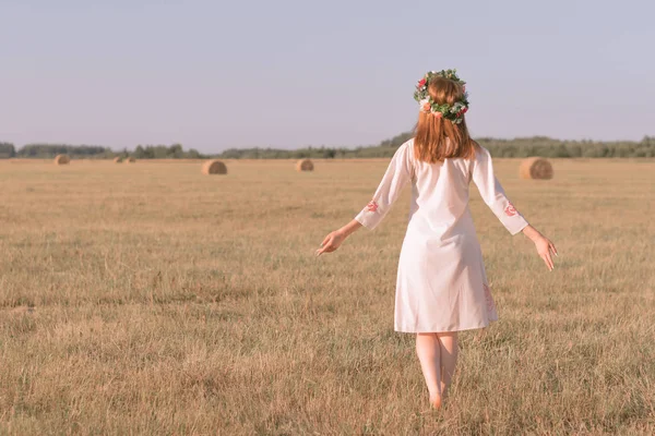 A young girl with red hair, standing alone in a field with grass in the summer, against the sky, forest and haystacks, in a sundress and with a wreath on his head, walking barefoot with his hands up.