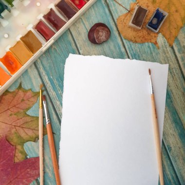 Creativity, objects for painting with copy space. A painted wooden table on which everything is prepared for drawing: a sheet of watercolor paper, watercolor paints, brushes, dry autumn leaves and chestnuts. 