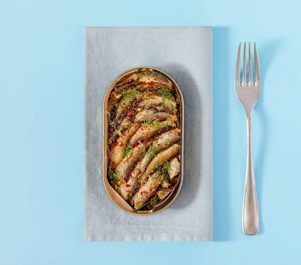 Open tin of Baltic sprats in oil with spices with a fork on a blue background. Snack from sea smoked fish. The concept of simple healthy food. Food photos, top view