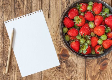 Bir kase olgun bektaşi üzümü, ahşap arka planda çilek, beyaz kağıt ve kalem.