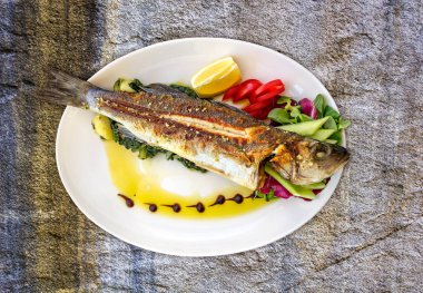 Taze otlu kızarmış balık, limon ve sebzeler beyaz tabakta, doğal taştan bir zemin üzerinde.