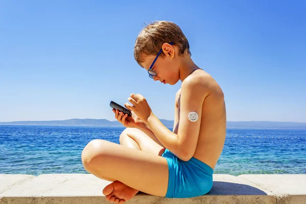 Summer sea holidays: a boy, a schoolboy sitting in a Lotus position on the beach measuring sugar levels using a sugar monitoring system. Concept life of a child with diabetes, glycemic control
