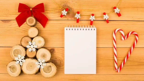 Noel ağacı ahşap kesim bir kırmızı yay ve beyaz kar taneleri ile yapılmış. Bunun yanında notlar için beyaz bir dizüstü bilgisayardır. Bu seçeneğin yanında bir kalp şekerler yapılan işte. Üzerinde kar taneleri ile çelenk, şurada. Her şey bir ahşap arka plan üzerinde olduğunu.
