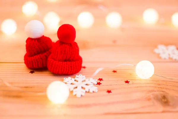 Kırmızı şapka ve ışıklar. Ponpon ile Winter.Two kırmızı şapka. Yakınlarda bir beyaz tahta kar tanesi var. Çelenk ışık onları çevreleyen. Tüm bu ahşap bir arka plan üzerinde.