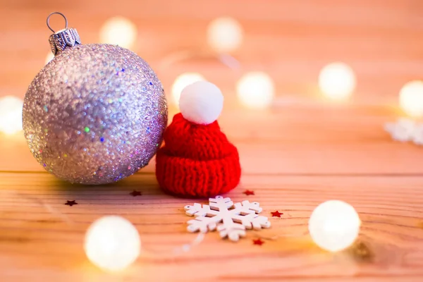 Gümüş bir Noel top ve bir beyaz kar tanesi yanında kırmızı bere. Çelenk ışık onları çevreleyen. Tüm bu ahşap bir arka plan üzerinde.