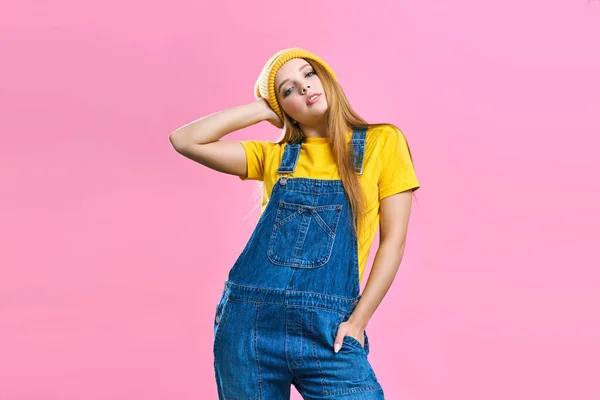 A pretty girl in denim overalls and a yellow hat on a pink background. Fashionista lady student. Bright trendy studio fashion image of sexy model, wearing neon bright color block clothes, casual