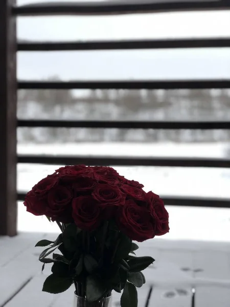 red roses in a vase on a snow-covered wooden brown balcony in a country house
