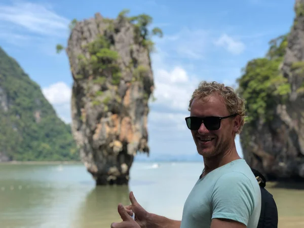 a light-skinned guy in sunglasses and a t-shirt shows his fingers the sign perfectly on the famous James bond island in Thailand, the azure sea and the wonder Islands in the afternoon